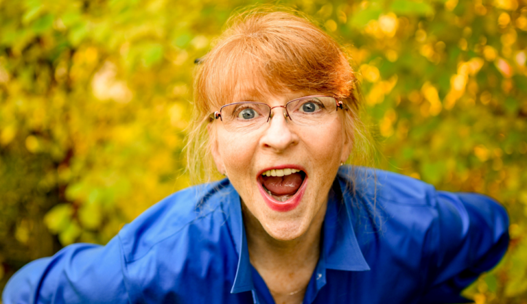 Woman with red hair and glasses wearing a blue shirt, smiling with her mouth open in a sunny, leafy outdoor setting.