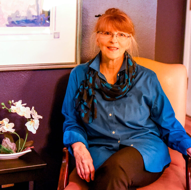 Smiling older woman with glasses in a blue blouse and teal scarf, seated indoors beside a purple wall.